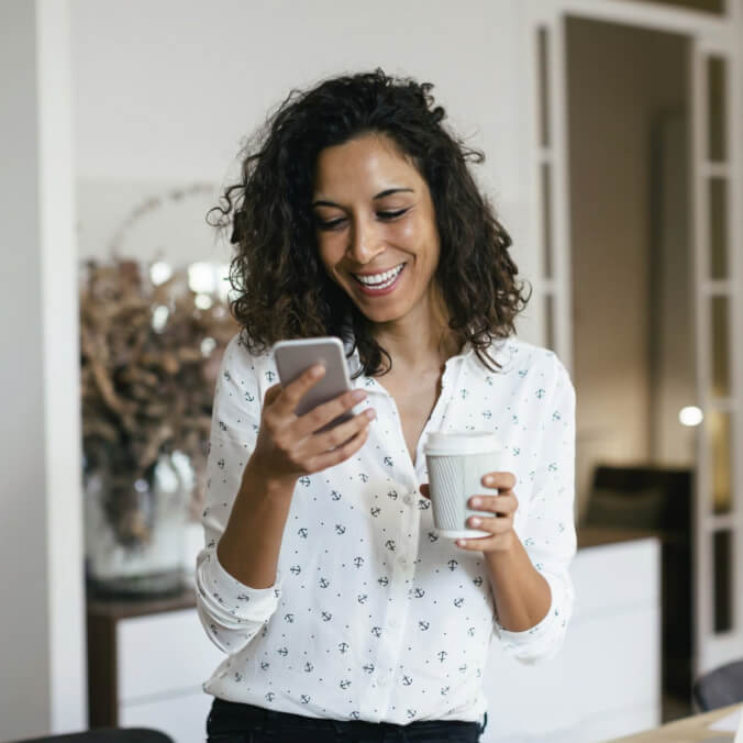 Una mujer sonriendo mientras ve su teléfono y sostiene un vaso de café.